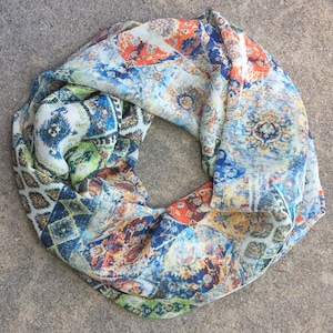 May include: A colorful infinity scarf with a vibrant, patterned design. The scarf features a mix of blue, orange, green, and white hues in a repeating, ornate pattern. The lightweight fabric is draped in a circular shape.