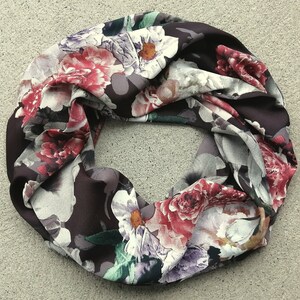 May include: A floral infinity scarf in a circular shape. The scarf has a dark burgundy base, adorned with large flowers in red, pink, white, and purple. The fabric appears soft and flowing, suitable for a variety of outfits.