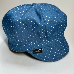 Peut inclure: Une casquette gavroche bleue ornée d'un motif à pois blancs sur tout le tissu. La casquette présente une visière courte et incurvée ainsi qu'une petite étiquette rectangulaire noire sur le bord avant avec le texte blanc Sanibel et une icône de palmier.