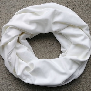 May include: A solid white infinity scarf made of a soft, smooth fabric. The scarf is arranged in a circular loop, featuring multiple folds and drapes that create a voluminous, layered appearance. The material has a matte finish and a clean, uniform colour.