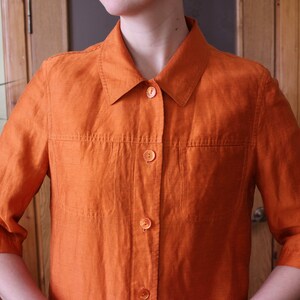 May include: A bright orange linen shirt with a collared neckline and button closure. The shirt has two pockets on the front and three-quarter sleeves.