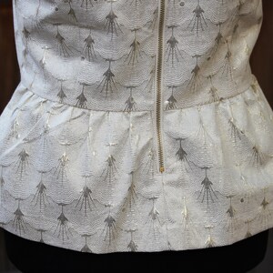 May include: A white top with a gold pattern and a zipper down the back. The top has a ruffled hem.