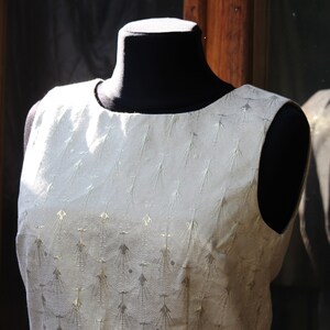 May include: A sleeveless dress with a white background and a gold pattern. The dress is on a black mannequin.
