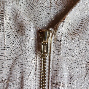 May include: Close-up of a gold zipper on a white fabric with a subtle metallic pattern.