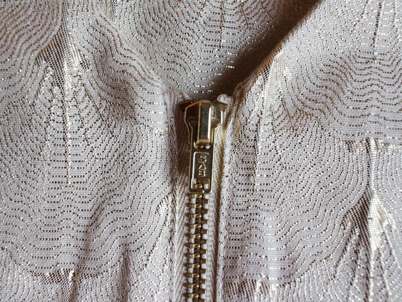 May include: Close-up of a gold zipper on a white fabric with a subtle metallic pattern.