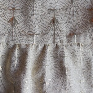 May include: Close-up of a white fabric with a silver metallic pattern. The fabric has a ruffled texture and a repeating design of curved lines.