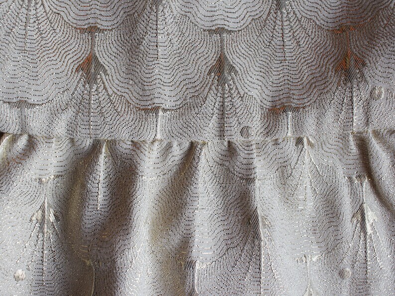 May include: Close-up of a white fabric with a silver metallic pattern. The fabric has a ruffled texture and a repeating design of curved lines.