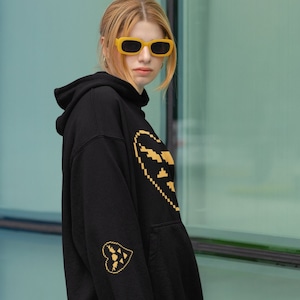 Puede incluir: Una sudadera con capucha negra con un diseño de corazón pixelado dorado en la parte delantera y un diseño de corazón pixelado dorado más pequeño en la manga izquierda. La modelo lleva gafas de sol amarillas.