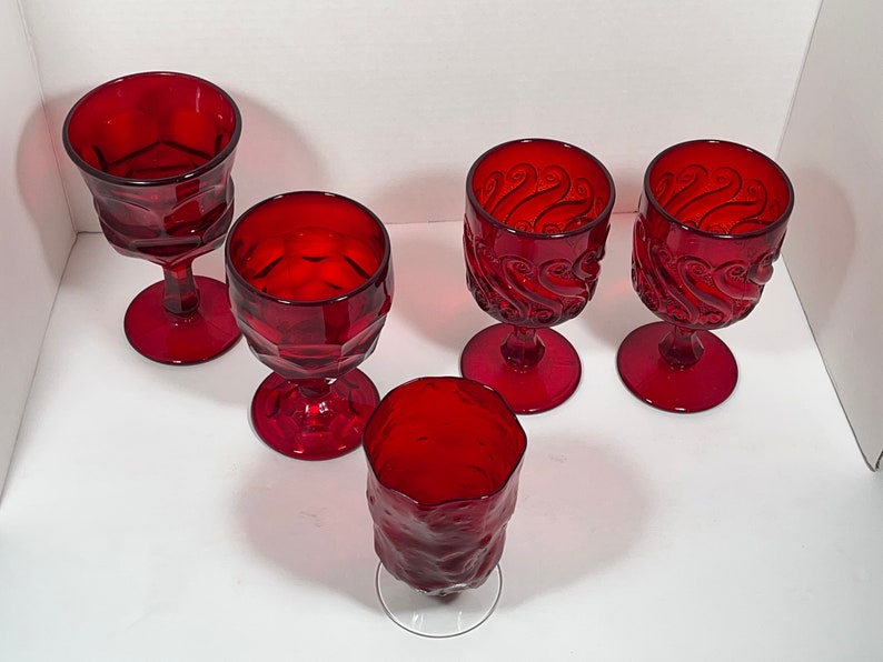 May include: A collection of five ruby red goblets. The glasses feature various designs, including geometric patterns and swirling textures. The goblets have a stem and a round base. The glassware is suitable for serving beverages.