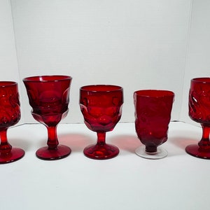 May include: Five ruby red vintage wine glasses in various styles. The glasses have ornate designs, including swirl and textured patterns. The stems and bases are also red, except for one with a clear base. The glasses are arranged on a white surface.