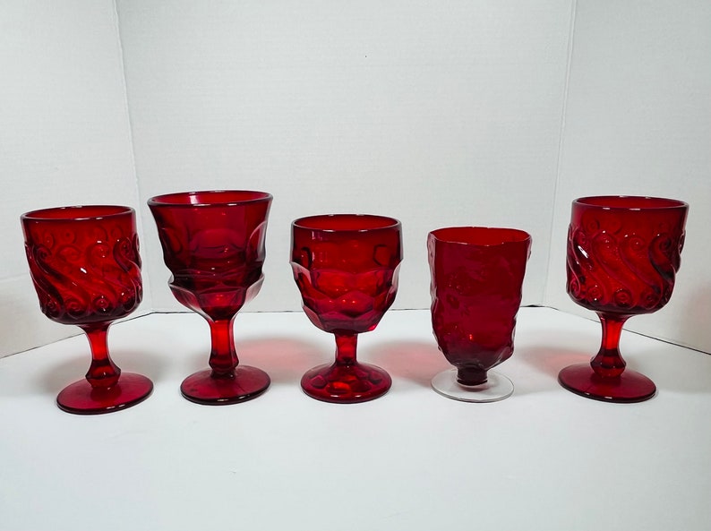 May include: Five ruby red vintage wine glasses in various styles. The glasses have ornate designs, including swirl and textured patterns. The stems and bases are also red, except for one with a clear base. The glasses are arranged on a white surface.