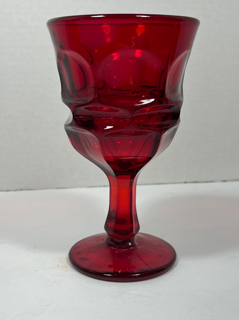 May include: A ruby red glass goblet with a textured, geometric design. The stem and base are solid red, and the bowl has a series of rounded, raised patterns. The glass has a high-gloss finish.