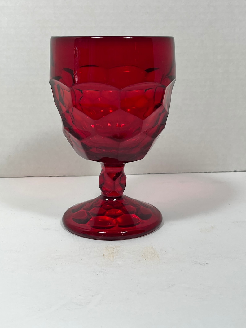 May include: A ruby red goblet with a faceted design. The glass has a stem and a round base. The goblet is made of glass and is suitable for serving beverages. The glass is empty.