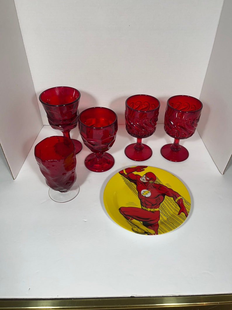 May include: A collection of red glassware and a yellow plate featuring The Flash. The glassware includes five wine glasses with varying designs. The plate has a red and yellow color scheme with a graphic of The Flash.