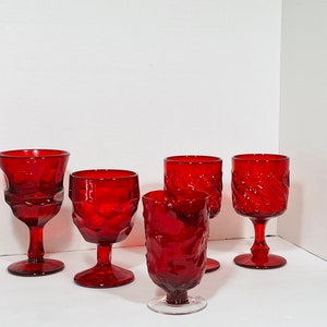 May include: A collection of six ruby red vintage wine glasses. The glasses feature various textured designs, including geometric patterns and swirling textures. Each glass has a stem and a bowl for holding liquid.