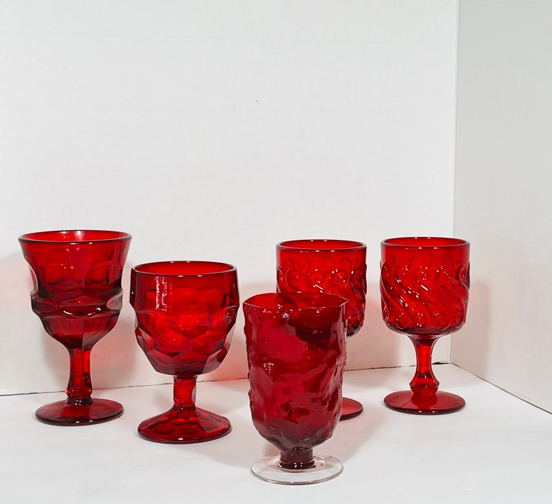 May include: A collection of six ruby red vintage wine glasses. The glasses feature various textured designs, including geometric patterns and swirling textures. Each glass has a stem and a bowl for holding liquid.