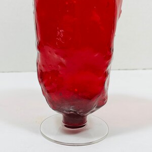 May include: A tall, red glass with a textured surface and a clear, circular base. The glass has a unique, organic shape and is likely handmade. The vibrant red color adds a striking visual element to the glassware.