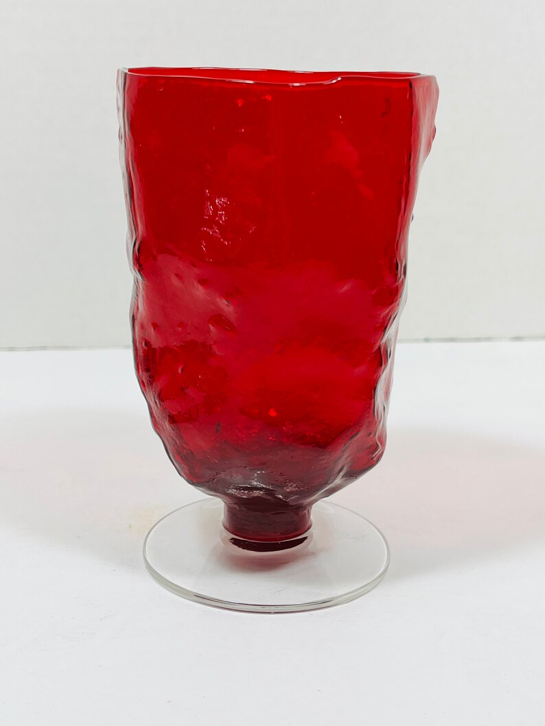 May include: A tall, red glass with a textured surface and a clear, circular base. The glass has a unique, organic shape and is likely handmade. The vibrant red color adds a striking visual element to the glassware.
