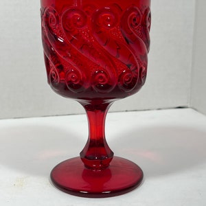 May include: A ruby red glass goblet with a decorative swirl pattern. The goblet has a round base, a slender stem, and a wide bowl. The glass is transparent, allowing light to pass through. The goblet is likely vintage or antique.