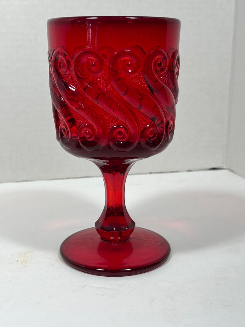 May include: A ruby red glass goblet with a decorative swirl pattern. The goblet has a round base, a slender stem, and a wide bowl. The glass is transparent, allowing light to pass through. The goblet is likely vintage or antique.