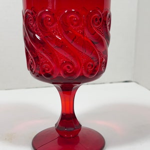 May include: A ruby red goblet with a detailed, swirling pattern around the bowl. The stem and base are also red, creating a cohesive and elegant design. The glass has a vintage aesthetic.