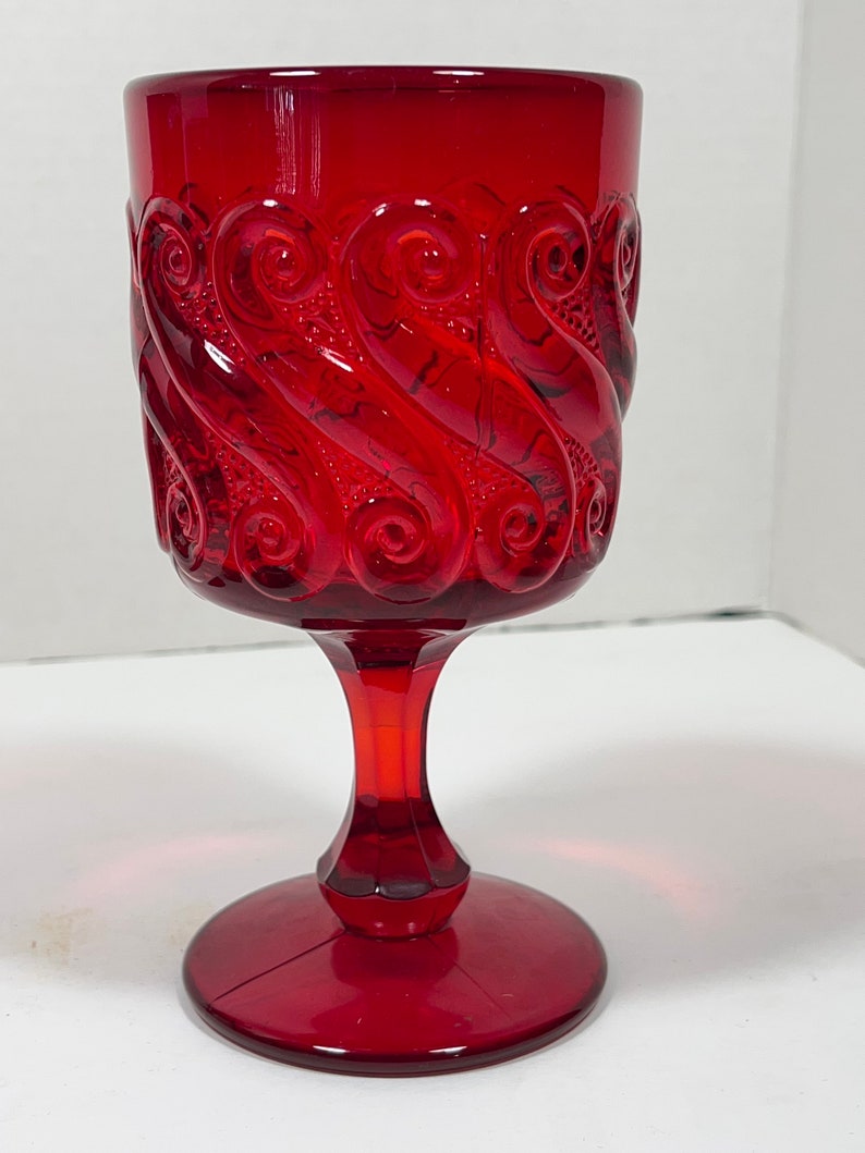 May include: A ruby red goblet with a detailed, swirling pattern around the bowl. The stem and base are also red, creating a cohesive and elegant design. The glass has a vintage aesthetic.