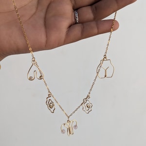 Puede incluir: Collar de oro con múltiples dijes, incluyendo un corazón, dos flores estilizadas, un par de senos, un par de caderas y un útero con dos cuentas rosadas.