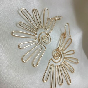 Puede incluir: Un par de pendientes de color dorado con un diseño en espiral y un diseño en forma de abanico. Los pendientes están hechos de alambre y tienen un aspecto delicado y femenino.