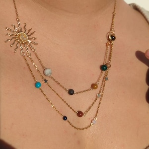 Puede incluir: Un collar de cadena dorada con un dije de sol y pequeñas cuentas que representan planetas. Las cuentas son de varios colores, incluyendo turquesa, marrón, verde y rojo.