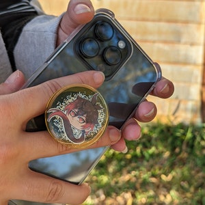 May include: A black smartphone with a clear case and a gold pop socket featuring a cartoon character. The character has red hair, black eyes, and is wearing a white shirt.