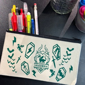 May include: A white canvas pencil case with a green dragon and the text "DIASOMNIA" printed on it. The case is decorated with green lightning bolts, bats, and swords.