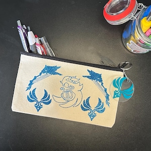 May include: A rectangular beige canvas pencil case with a black zipper closure. The front features blue glitter graphics of a central lyre emblem flanked by two stylized wing motifs and two diagonal arrow designs. A circular clear acrylic charm hangs from the zipper.