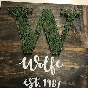 May include: A wooden sign with the letter W made from string art in green thread. The sign also has the text "Wolfe est. 1987" written in white paint.