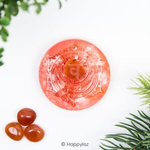 Puede incluir: Una placa de chakra de cristal rojo con un símbolo tallado y un cristal rojo más pequeño en la parte superior. Tres cristales rojos más pequeños se encuentran en la esquina inferior derecha de la imagen.
