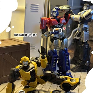 May include: A photograph featuring several Transformers action figures. The figures are in various poses, with Bumblebee on the ground and Optimus Prime holding a small can. The figures are primarily red, blue, yellow, and silver. The Etsy shop name "owlcatcrafts etsy" is visible.