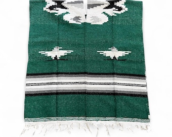 May include: A green woven poncho with a geometric design in black, white, and gray. The poncho features a central opening for the head and a fringed bottom edge. The design includes bird-like shapes and horizontal stripes.