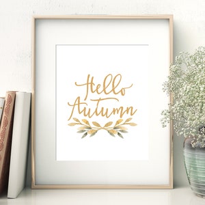 May include: A framed print with the text "Hello Autumn" in gold script lettering. The text is surrounded by a wreath of watercolor leaves in shades of green and gold.