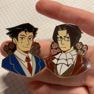 May include: Two enamel pins featuring anime-style characters. The left pin shows a person with dark hair in a blue suit and red tie. The right pin shows a person with dark hair and glasses in a brown suit and white cravat. Both pins feature red roses.