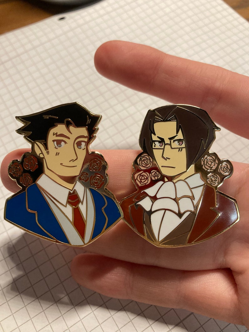 May include: Two enamel pins featuring anime-style characters. The left pin shows a person with dark hair in a blue suit and red tie. The right pin shows a person with dark hair and glasses in a brown suit and white cravat. Both pins feature red roses.