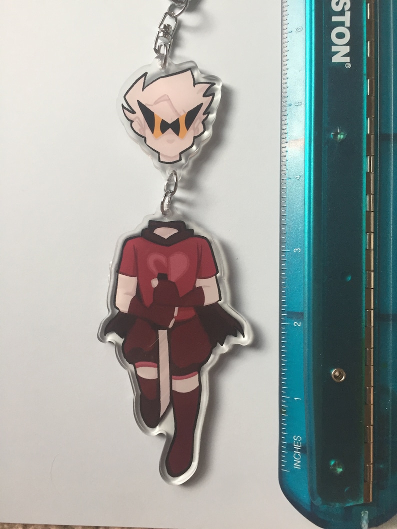 May include: A clear acrylic keychain featuring two cartoon character designs. The top design is a head with light pink hair and black and orange glasses. The bottom design is a red and maroon outfit with a sword. The keychain is attached to a silver chain.
