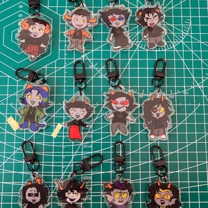 May include: Collection of acrylic keychains featuring cartoon characters with horns and various outfits. Each keychain has a black clasp. The characters have different skin tones, hair colors, and expressions. The keychains are displayed on a green cutting mat.