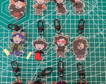 May include: Collection of acrylic keychains featuring cartoon characters with horns and various outfits. Each keychain has a black clasp. The characters have different skin tones, hair colors, and expressions. The keychains are displayed on a green cutting mat.