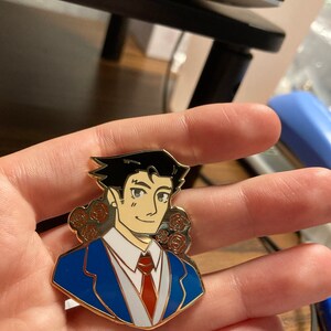 May include: A gold-toned metal enamel pin featuring a stylized illustration of a character with black hair, wearing a blue suit jacket, white shirt, and red tie. The design includes small red rose accents on both sides of the character's head.