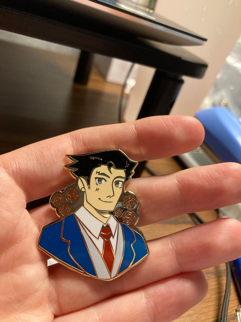 May include: A gold-toned metal enamel pin featuring a stylized illustration of a character with black hair, wearing a blue suit jacket, white shirt, and red tie. The design includes small red rose accents on both sides of the character's head.