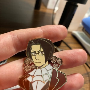 May include: A metal enamel pin featuring a stylized illustration of a character with dark brown hair, glasses, and a white cravat, wearing a dark brown suit jacket. The character is framed by small, light-colored rose motifs on both sides.