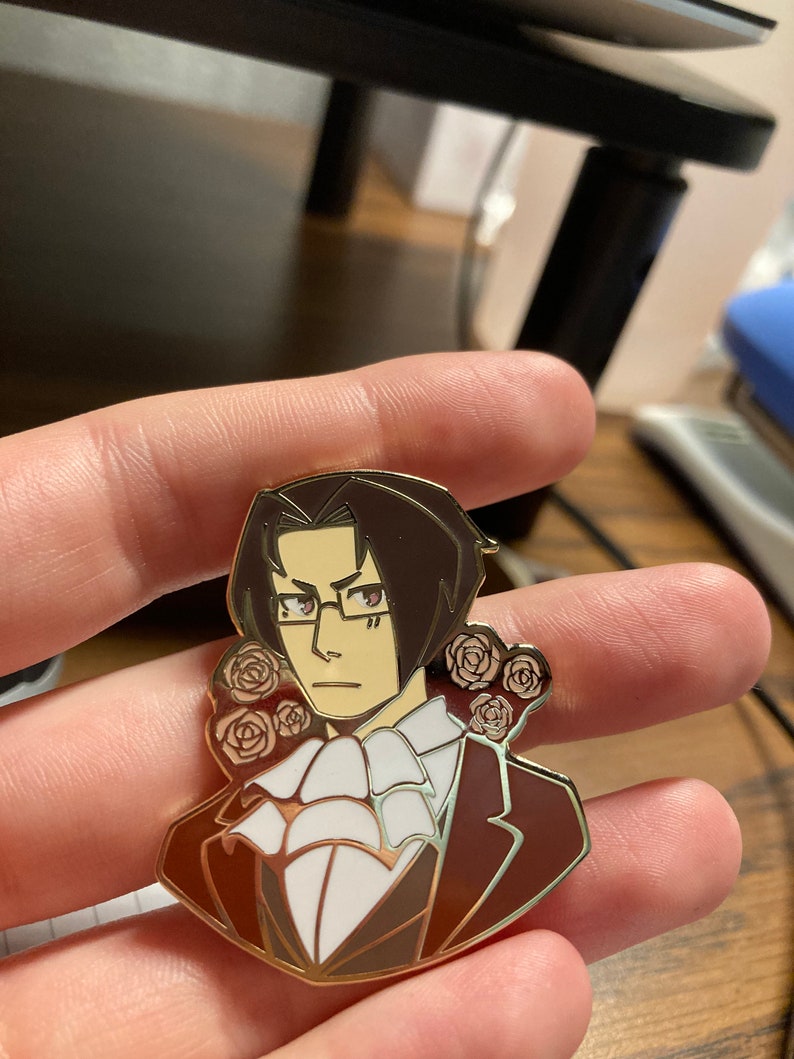 May include: A metal enamel pin featuring a stylized illustration of a character with dark brown hair, glasses, and a white cravat, wearing a dark brown suit jacket. The character is framed by small, light-colored rose motifs on both sides.