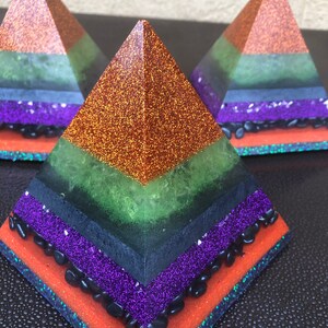 Könnte beinhalten: Drei mehrschichtige Harzpyramiden mit Glitzer und schwarzen Steinen. Die Pyramiden sind orange, grün, schwarz und lila.
