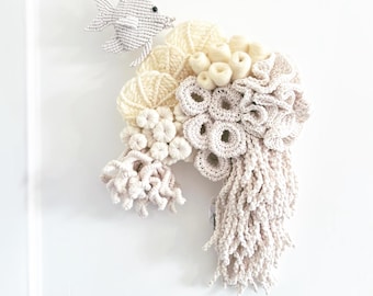 May include: A handmade macrame wall hanging with a fish and coral reef design. The artwork is mainly cream and off-white, with a driftwood accent. The fish is grey and hangs from a string. The reef has various textures and shapes.