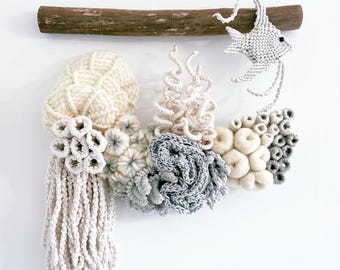 May include: A handmade wall hanging with a marine theme. The artwork features textured elements, including a cream-coloured jellyfish, coral, and a fish, all made from yarn. The piece is mounted on a wooden branch.
