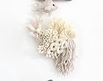May include: A macrame wall hanging depicting a fish and coral reef. The white fish with a black eye is suspended above a textured reef in cream and beige. The artwork is mounted on driftwood. The overall design evokes an underwater scene.